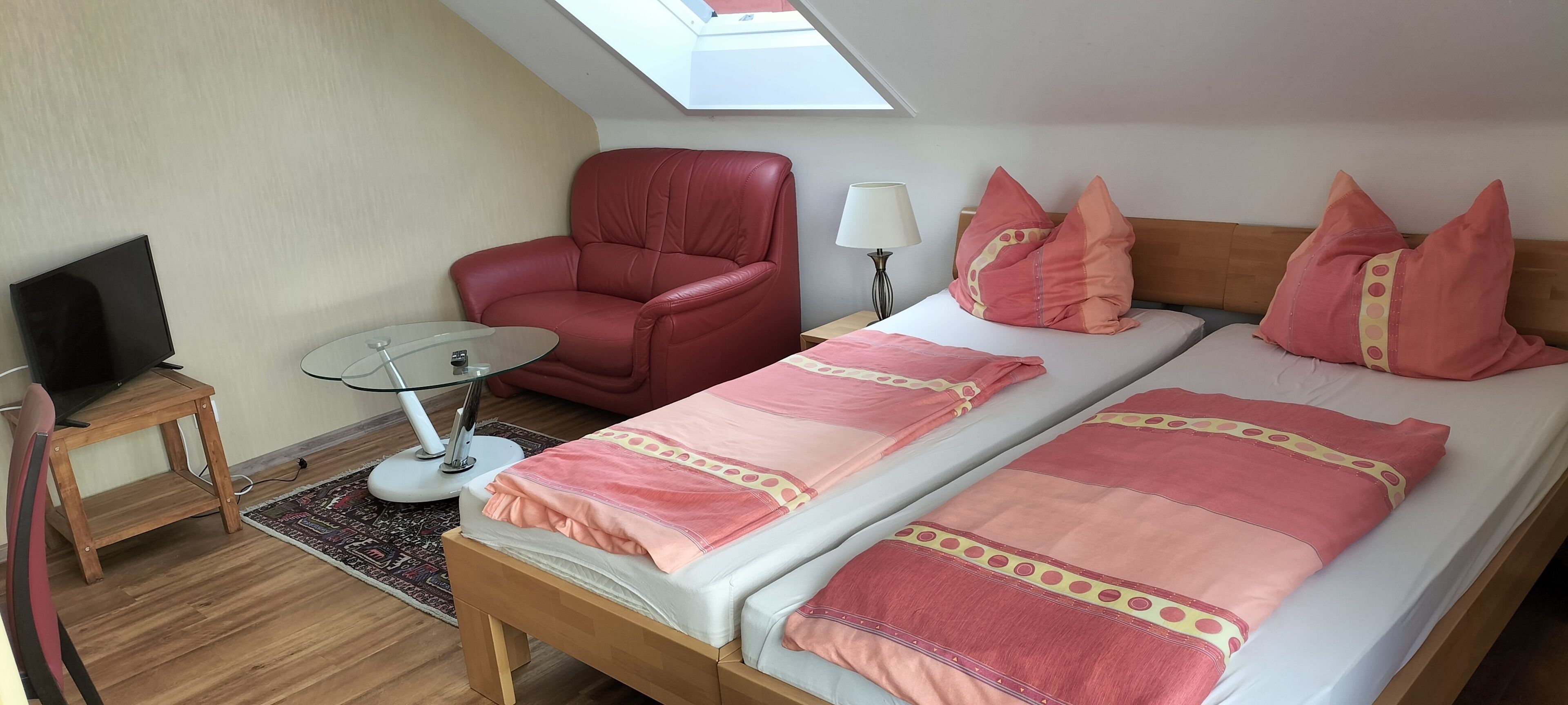 A room with a bed, a red chair, a small table with a lamp, and a TV.