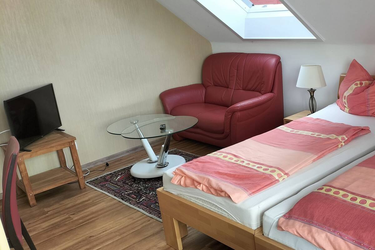 A room with a bed, a red chair, a small table with a lamp, and a TV.