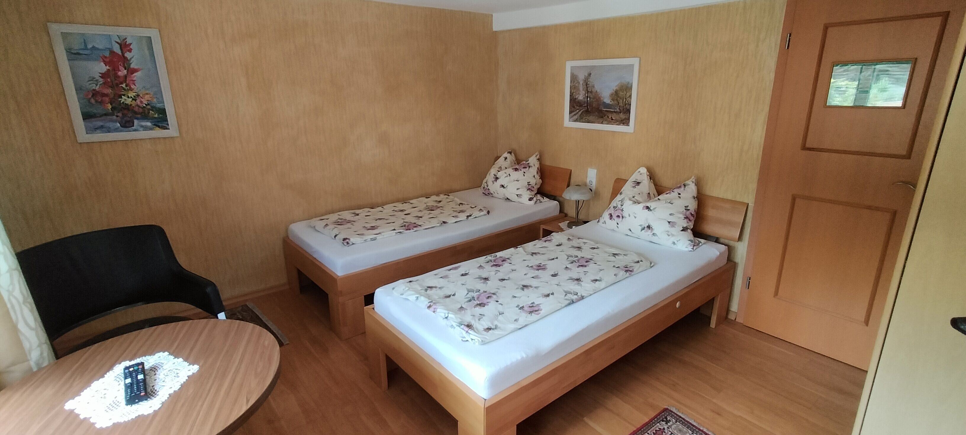Una habitación con dos camas individuales, una silla de madera, una mesita redonda con un control remoto y dos cuadros en las paredes.
