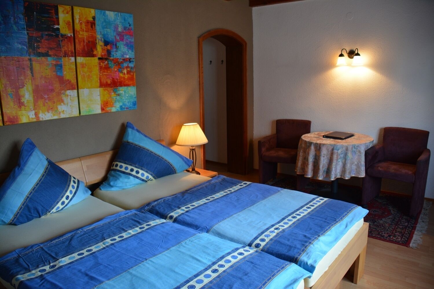 Una cama con ropa de cama azul, una lámpara, dos sillas, una mesita pequeña y un cuadro abstracto colorido en la pared.