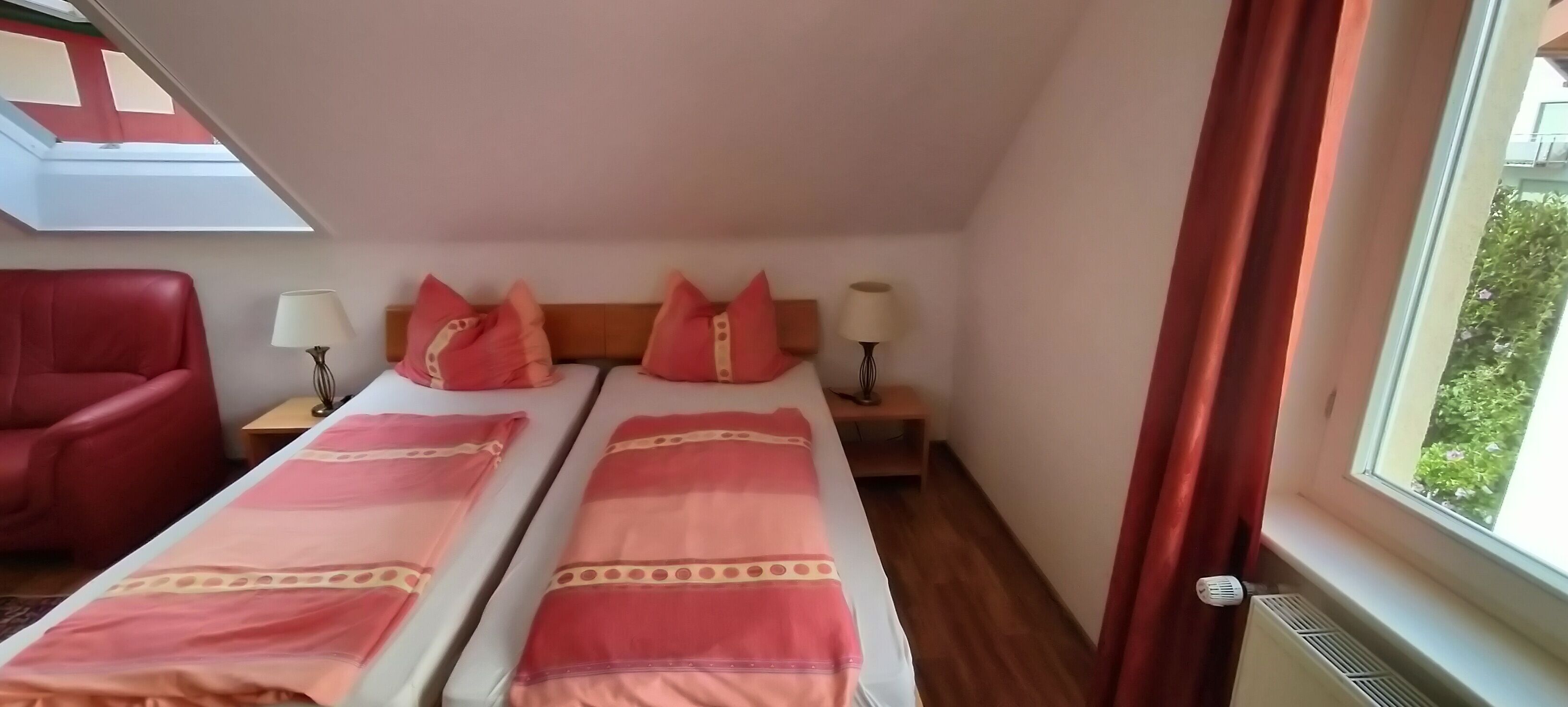 Habitación de hotel con dos camas, una silla roja, mesitas de noche con lámparas, un tragaluz y vistas a zonas verdes.