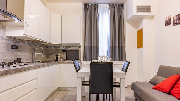 Uma cozinha compacta com armários brancos, uma mesa de jantar com cadeiras, um sofá cinza e uma janela com cortinas cinzas.