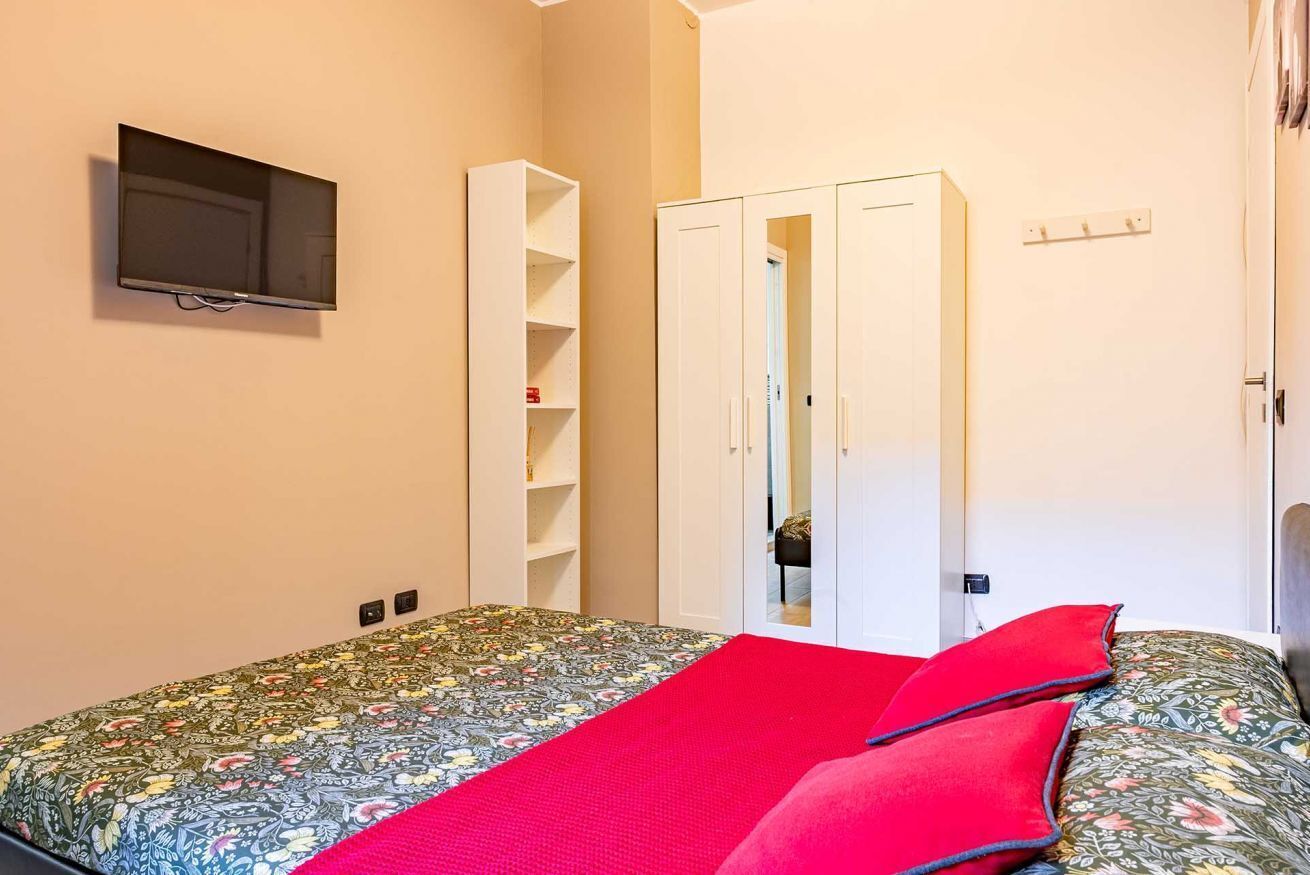 A bedroom with a bed, a TV mounted on the wall, a wardrobe, and a door leading to another room.