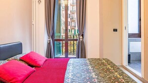 Um quarto com cama, janela com cortinas, vista para uma varanda e porta que dá para um banheiro.