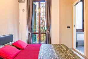 Ein Schlafzimmer mit Bett, einem Fenster mit Vorhängen, Blick auf einen Balkon und einer Tür zu einem Badezimmer.