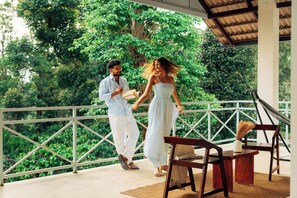 A couple walking on a balcony with a view of lush greenery, a wooden table, and chairs.