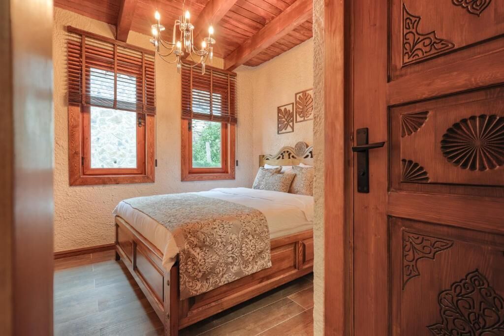 A bedroom with a wooden bed, two windows with shutters, a chandelier, and a wooden door with carvings.