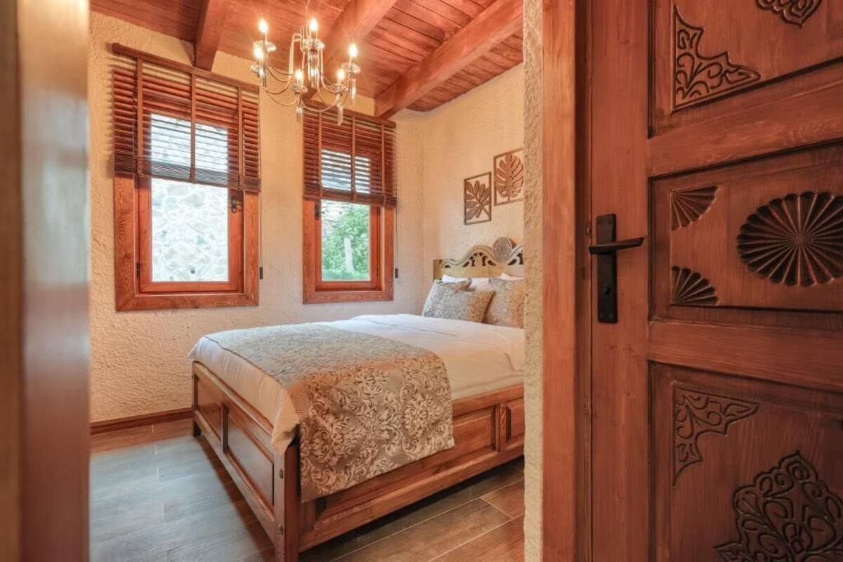 A bedroom with a wooden bed, two windows with shutters, a chandelier, and a wooden door with carvings.