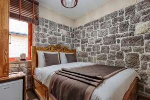 A room with a stone wall, a wooden bed with a headboard, a small kitchenette with a refrigerator, and a window with blinds. - KALE KONAK 17 (Eceabat)