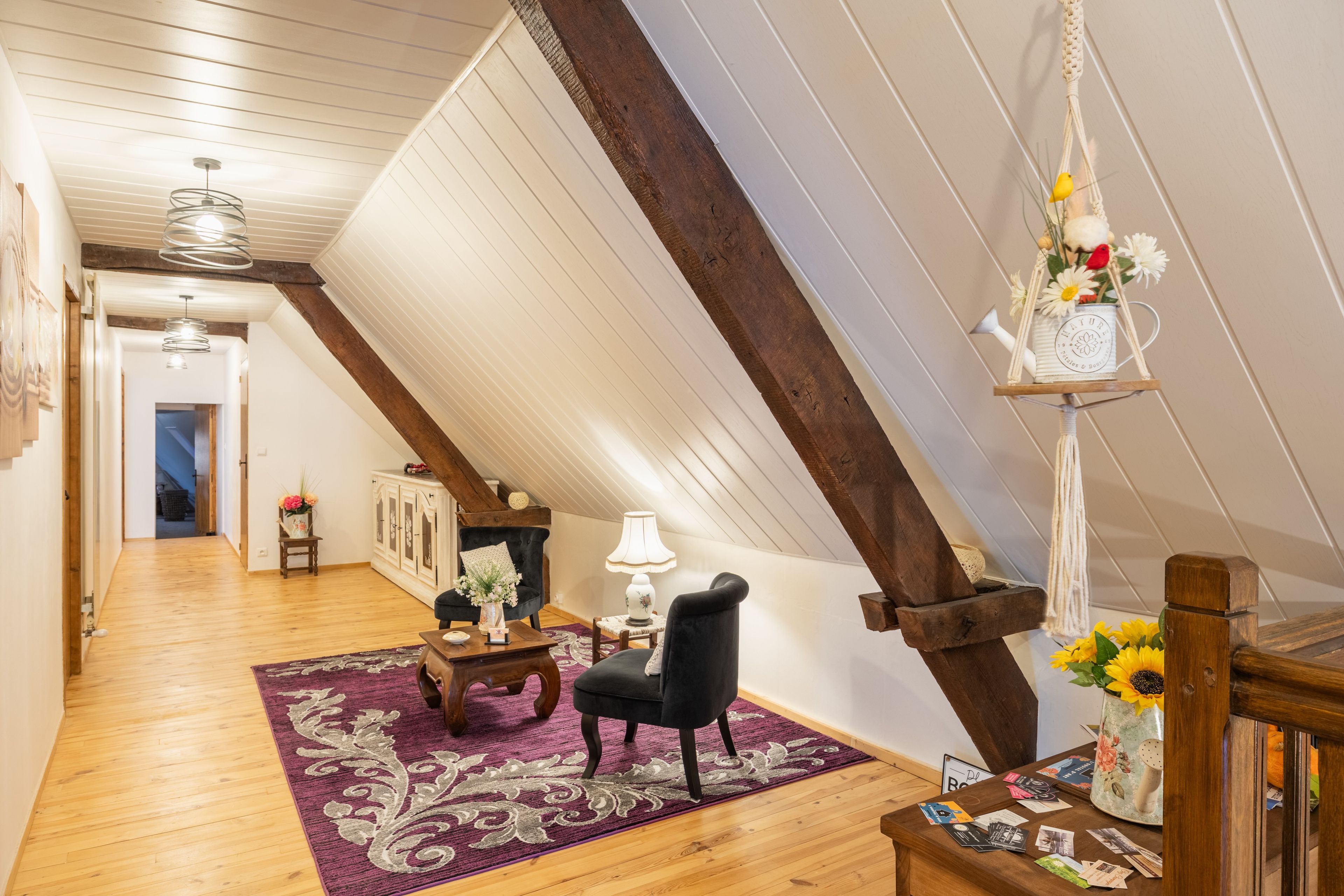 Ett rum i loftstil med synliga träbjälkar, en svart fåtölj, ett träbord och en hängande lampa i makramé.
