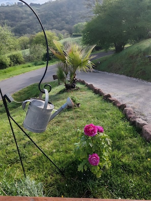 A garden with a watering can, a flower, and a road. - Sans Chichi Chambres et Table: Mountain View, Shared Terrace & Wi-Fi (Escandolières)