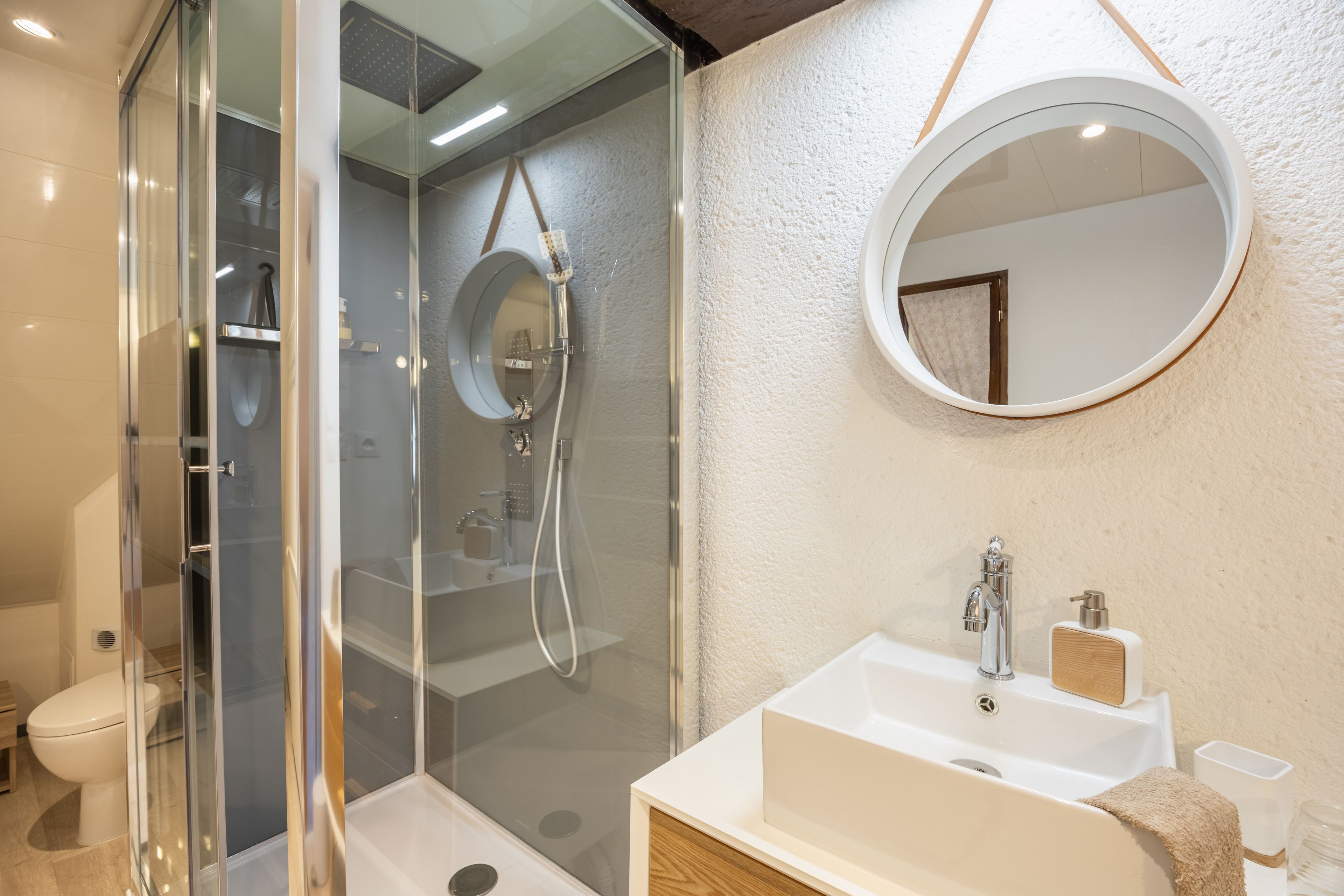 Une salle de bain moderne avec une douche à l’italienne en verre, un lavabo blanc avec un meuble-vasque en bois et un miroir rond au mur.