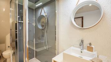 Une salle de bain moderne avec une douche à l’italienne en verre, un lavabo blanc avec un meuble-vasque en bois et un miroir rond au mur.