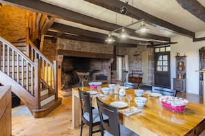 Un coin repas avec une table en bois dressée pour quatre personnes, entourée de chaises, dans une pièce aux poutres apparentes et aux murs en pierre.