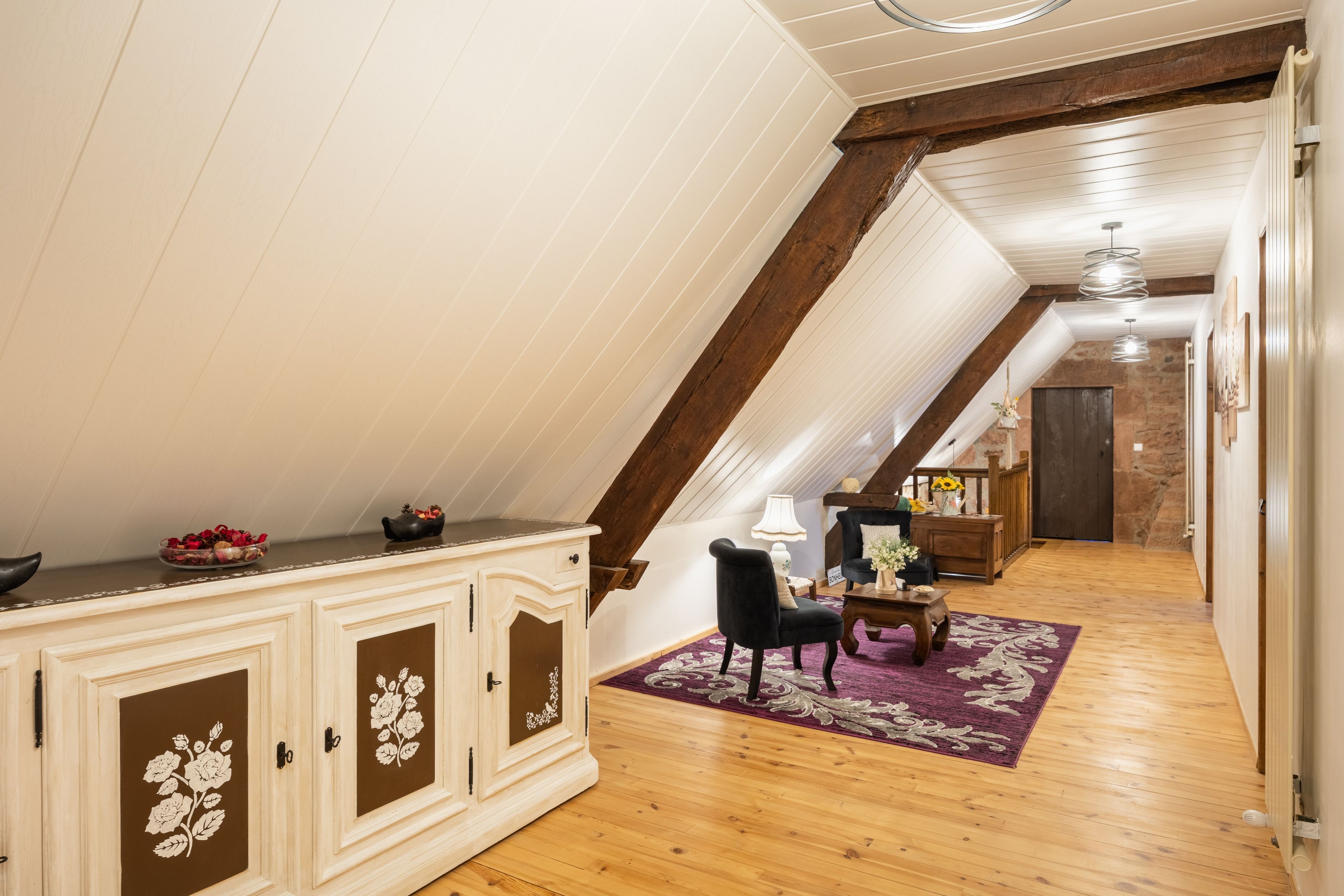 Een kamer met een schuine plafond, houten balken, een witte kast met bloemmotieven, een zwarte stoel, een klein tafeltje met een lamp en een paars tapijt.