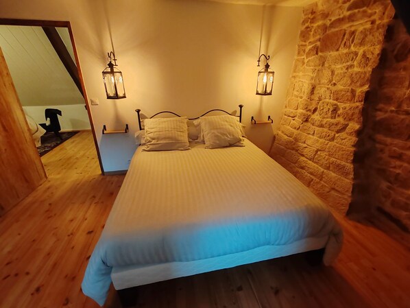 A bedroom with a wooden floor, a bed with white bedding, two wall-mounted lanterns, and a brick wall. - Sans Chichi Chambres et Table B&B with Terrace, Garden, and Wi-Fi (Escandolières)