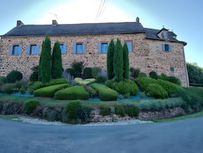 A stone building with a symmetrical facade, multiple windows with shutters, and a well-maintained garden with various shrubs and a small water feature.