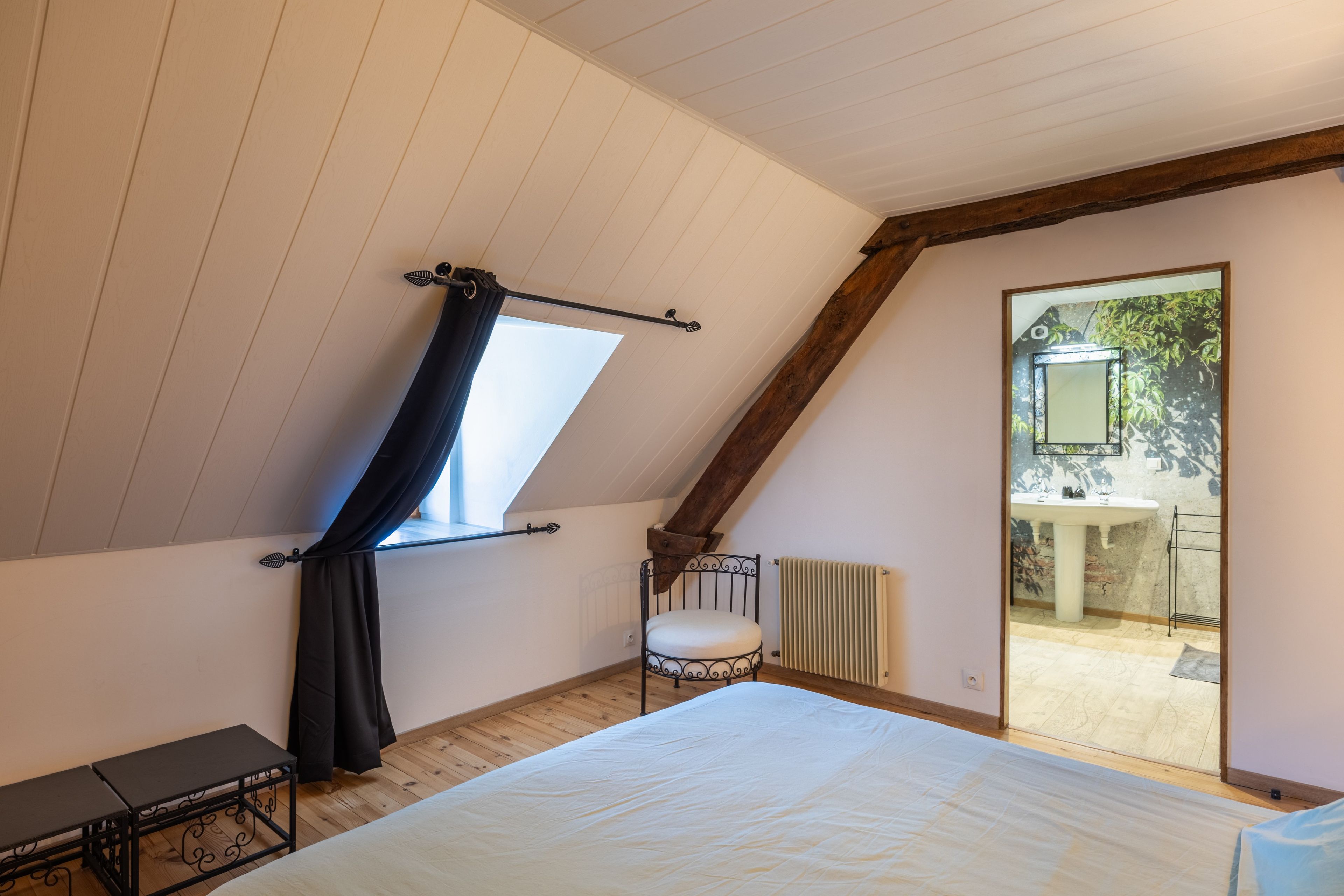 A bedroom with a sloped ceiling, a bed, a small table, a chair, and a window with curtains.