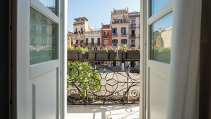 Vista a través de una ventana abierta con balcón, sobre una calle con edificios y un coche estacionado.