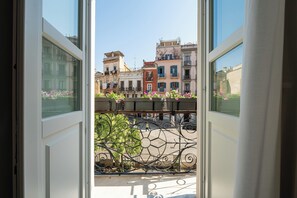 A view through an open window with a balcony, overlooking a street with buildings and a parked car.