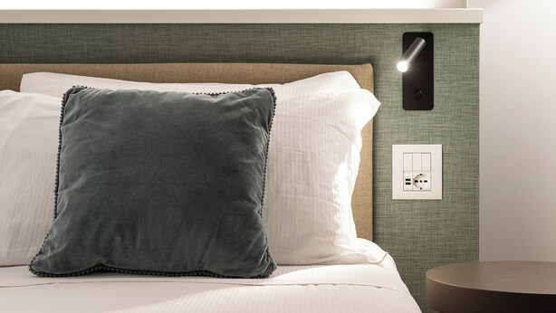 Una habitación de hotel con cama, almohadas, una lámpara fijada a la pared y un enchufe.