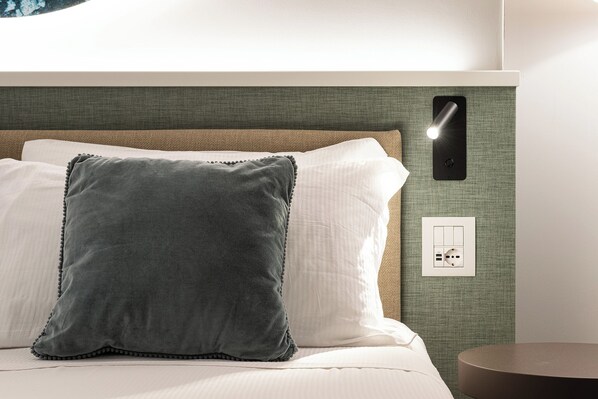 A hotel room with a bed, pillows, a wall-mounted light, and an electrical outlet.