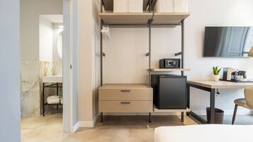 A compact bedroom with a bed, a desk with a computer, a small refrigerator, and a TV.