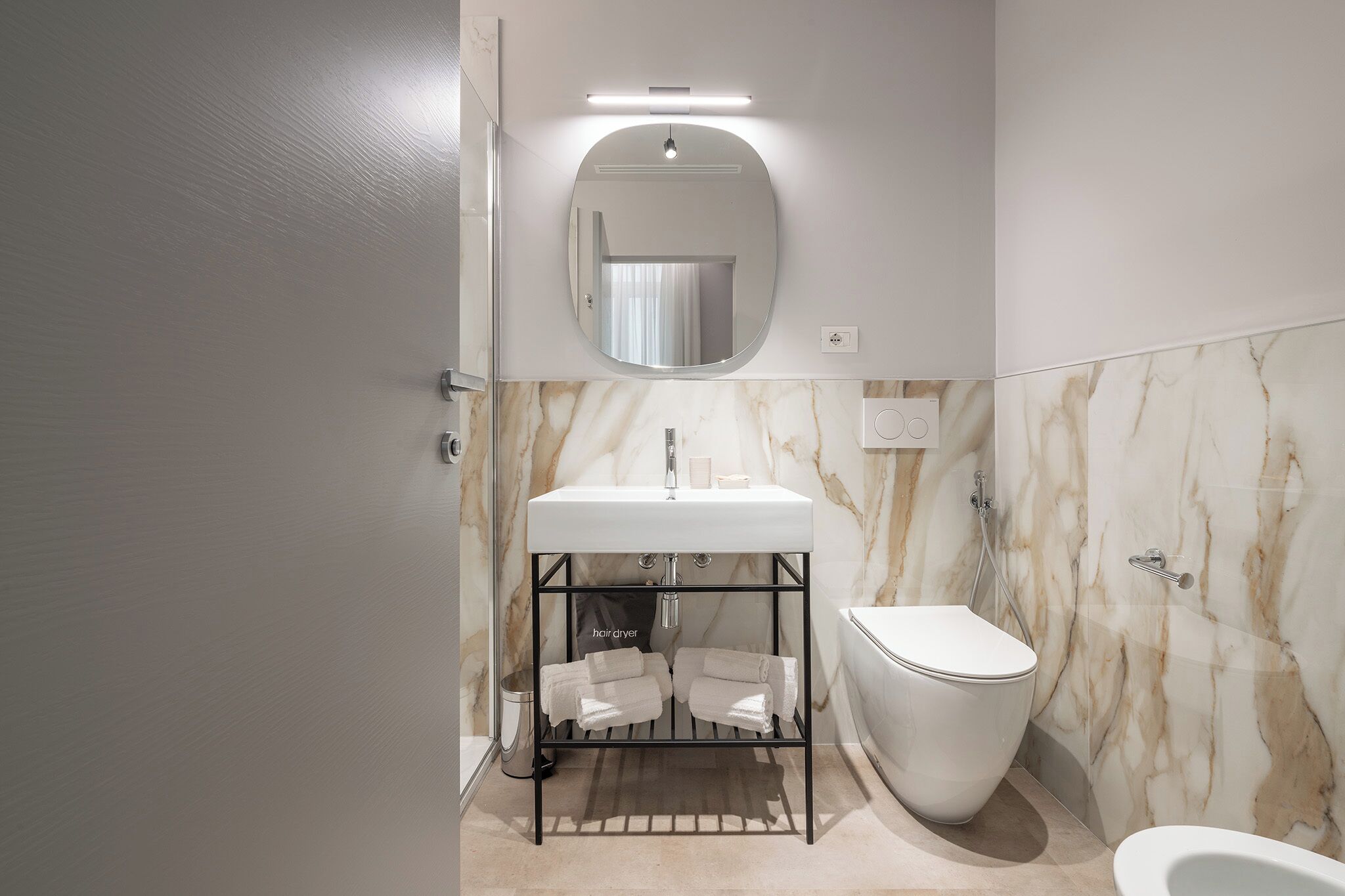 A modern bathroom with a marble wall, a white sink, a toilet, and a hair dryer on a rack.