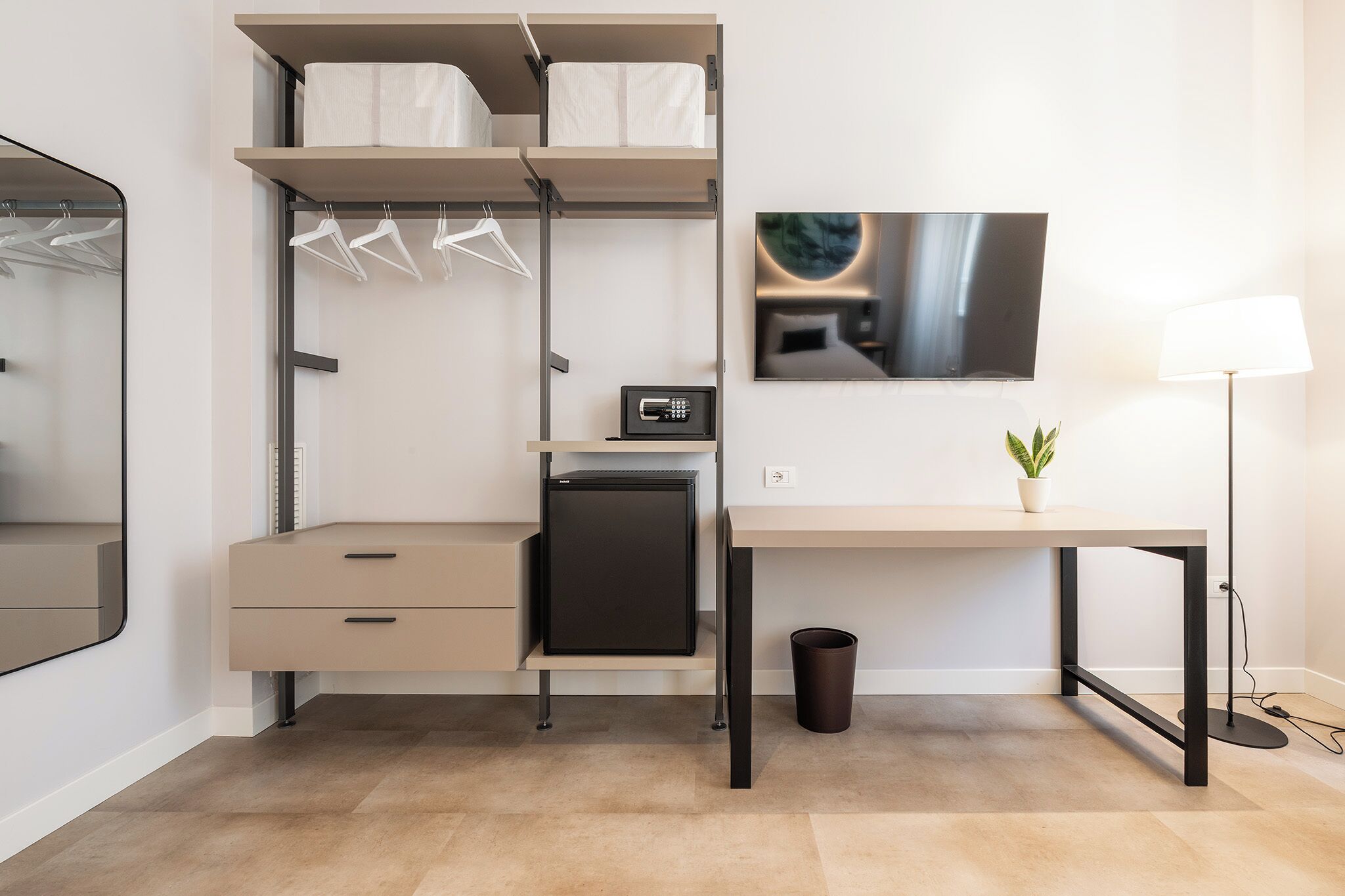 A compact bedroom with a bed, a desk, a TV, and a small plant.
