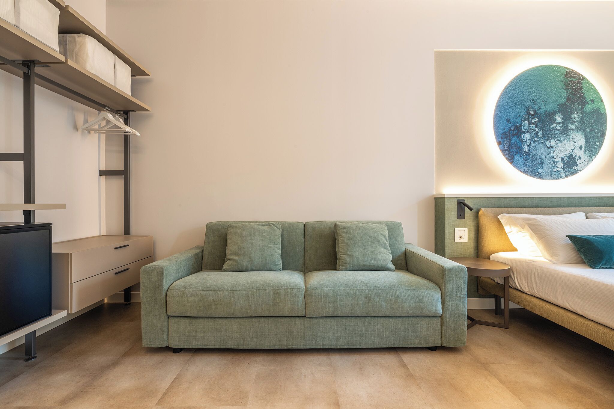 Un dormitorio moderno con un sofá verde, una cama con ropa de cama blanca, un televisor montado en la pared y una obra de arte circular sobre la cama.