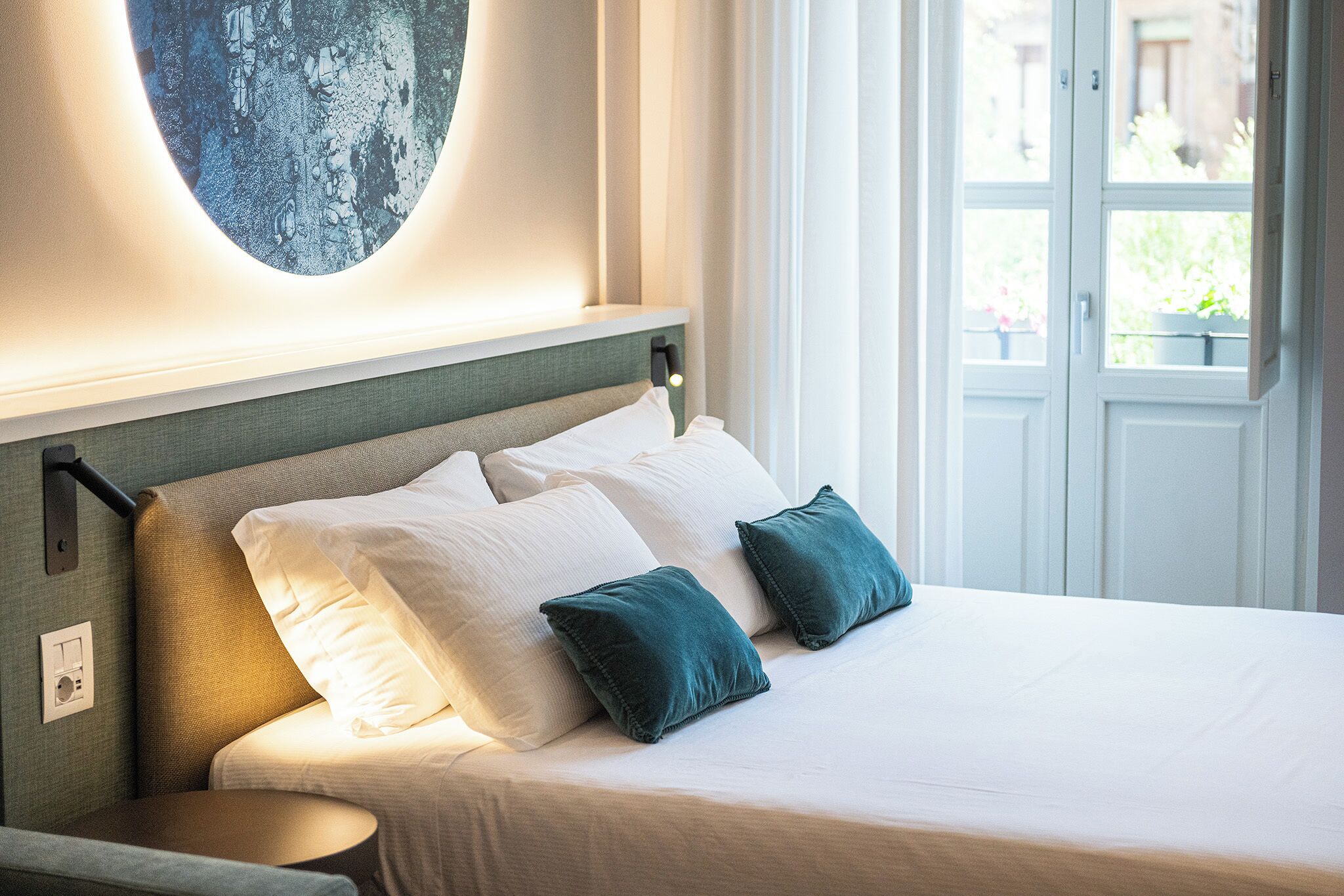 Un lit bien fait, agrémenté d’oreillers blancs et turquoise, une tête de lit au motif texturé et un luminaire fixé au mur.