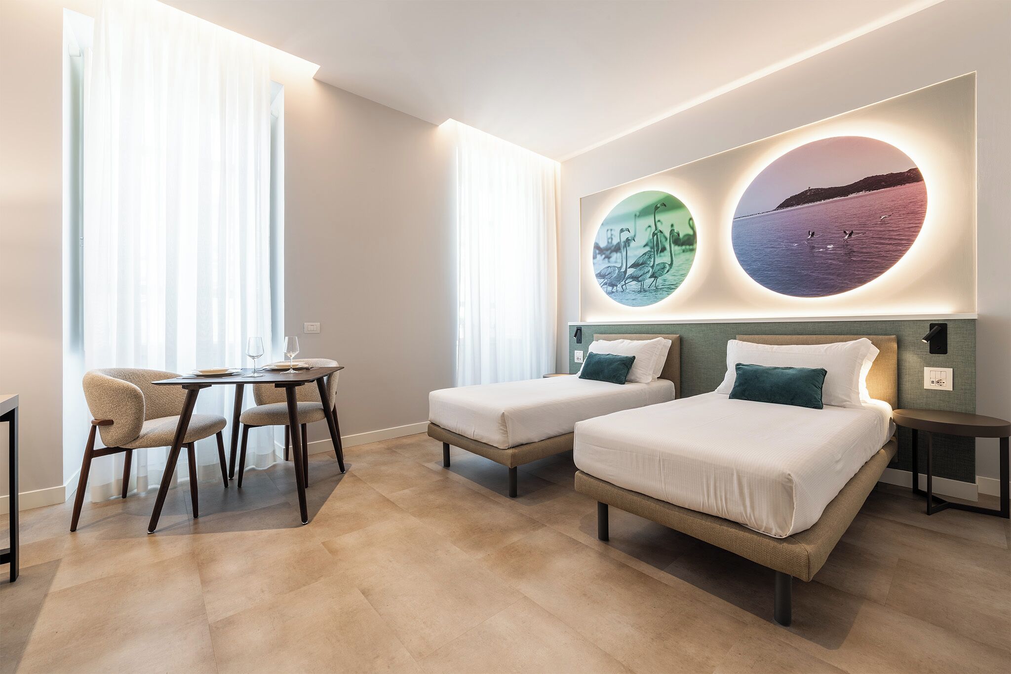 Ein Hotelzimmer mit zwei Betten, einem kleinen Tisch und Stühlen. An der Wand befinden sich zwei runde Leuchten mit Abbildungen.