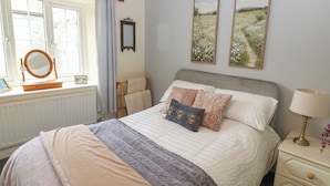 A bedroom with a bed, bedside table, lamp, and window with curtains. - Wedgewood Cottage (Leyburn)