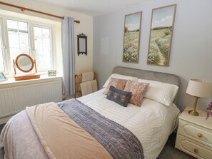 A bedroom with a bed, bedside table, lamp, and window with curtains. - Wedgewood Cottage (Leyburn)