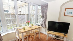 A room with a wooden dining table, chairs, a television on a stand, and a window with curtains. - Wedgewood Cottage (Leyburn)