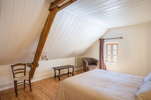 A room with a sloped ceiling, a bed, a chair, a small table, and a window with curtains.