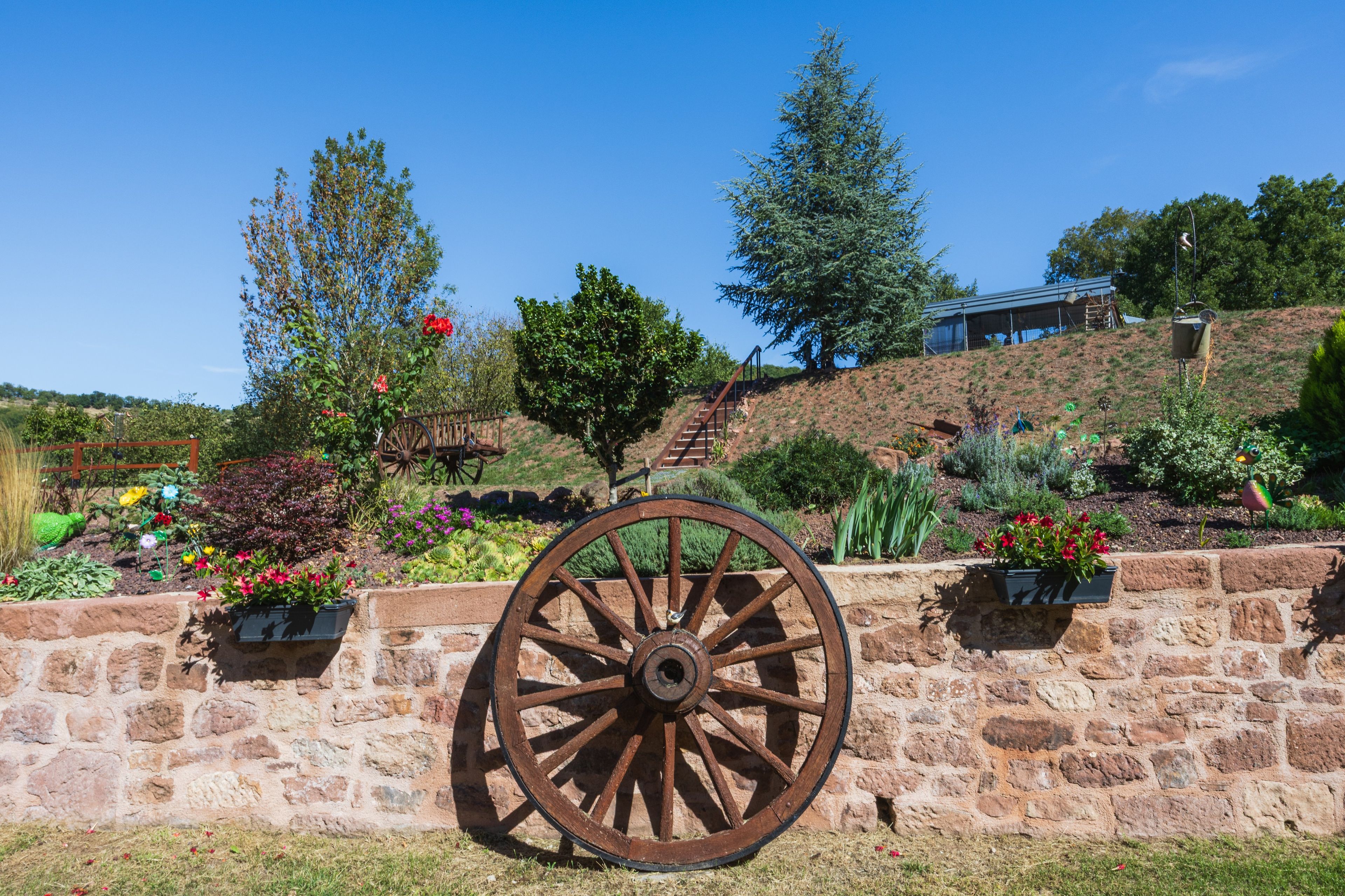 A stone wall with a large wooden wheel, surrounded by a variety of colorful flowers and plants.