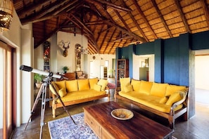 Ein Zimmer mit einem Strohdach, Holzbalken, einem Teleskop, gelben Sofas und einem Holztisch.