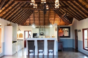 A spacious kitchen with a high ceiling, a central island with bar stools, and a dining area with a large mirror. - Parsons House (Hoedspruit)