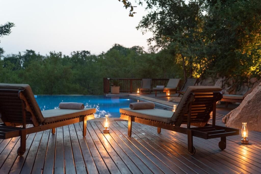Terraza de madera con sillones de descanso y velas, con vista a una piscina.