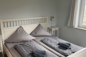 A neatly made bed with grey linens and pillows, a bedside table with a lamp, and a window with curtains. - Vacation Home "Sandbank" in Zierow on the Baltic Sea (Zierow)