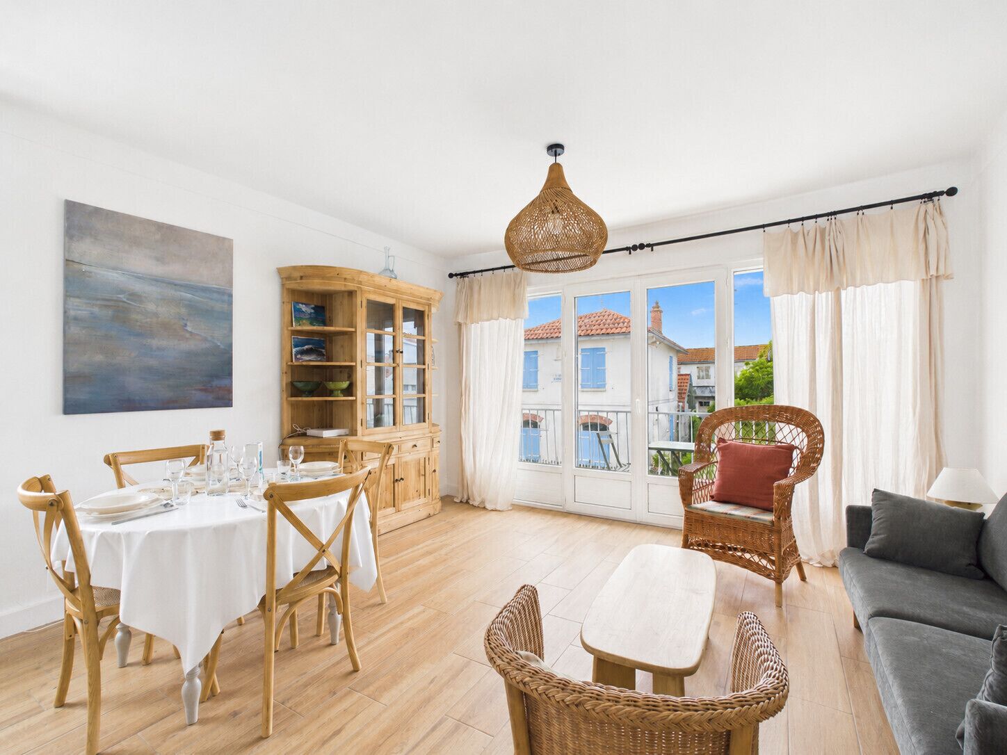 A dining area with a round table and chairs, a wicker sofa, a cabinet with glass doors, and a view through large windows.