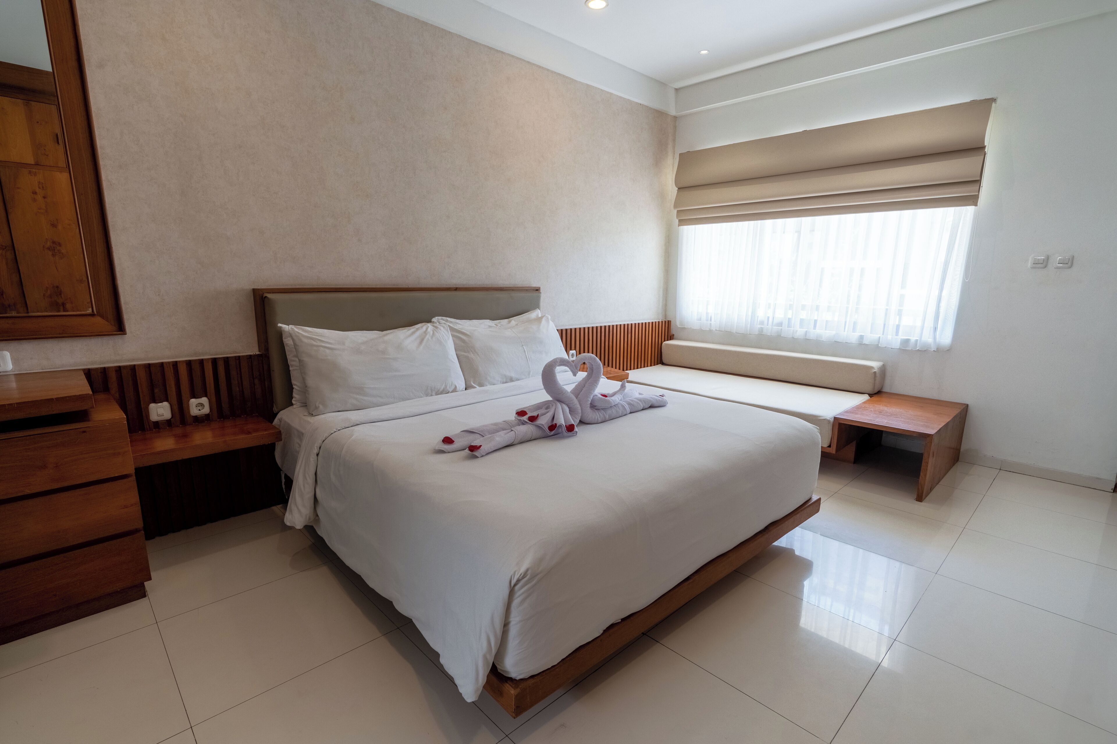 A hotel room with a large bed, a wooden nightstand, a wooden bench, a window with a blind, and a mirror on the wall.