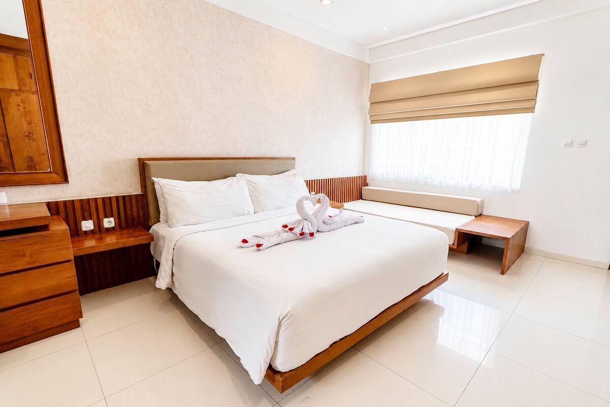 Una habitación de hotel moderna con una cama grande, mesitas de noche de madera, un banco y un ventanal con persianas.