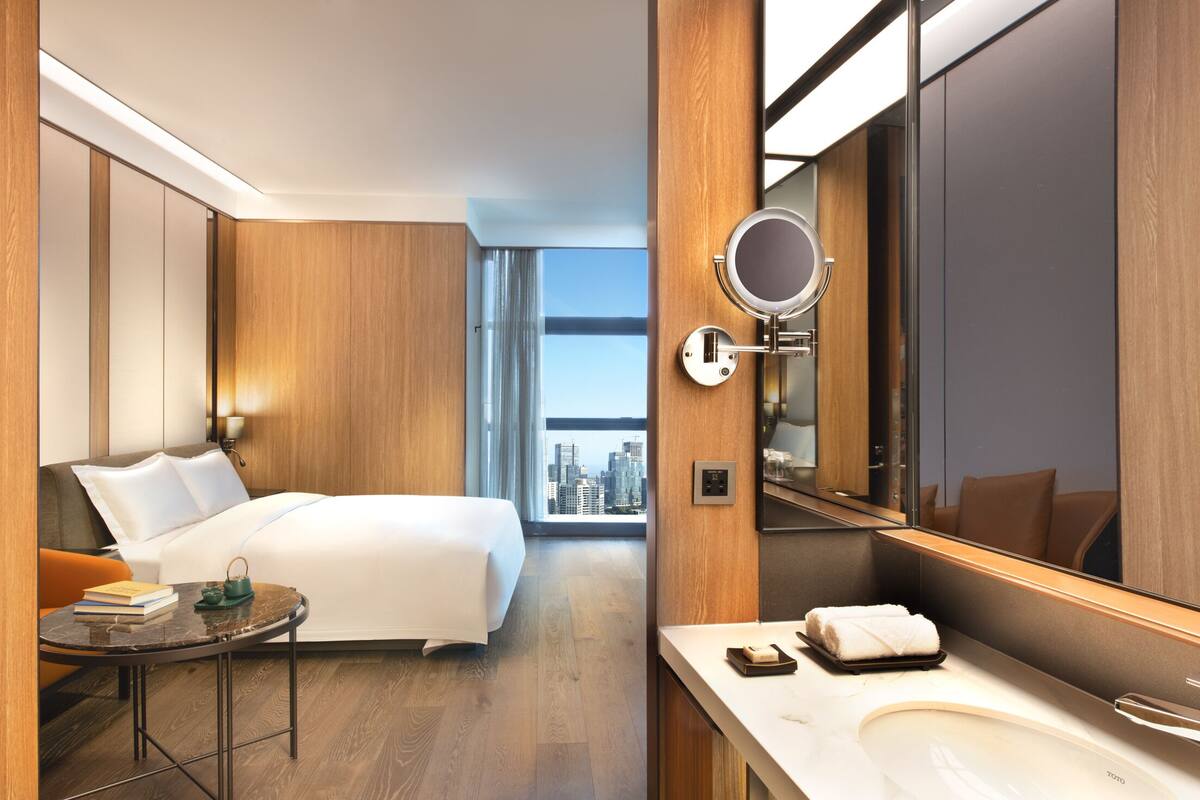 Habitación de hotel con cama, espejo redondo, una pequeña mesa con libros, baño con lavamanos y vista a la ciudad.