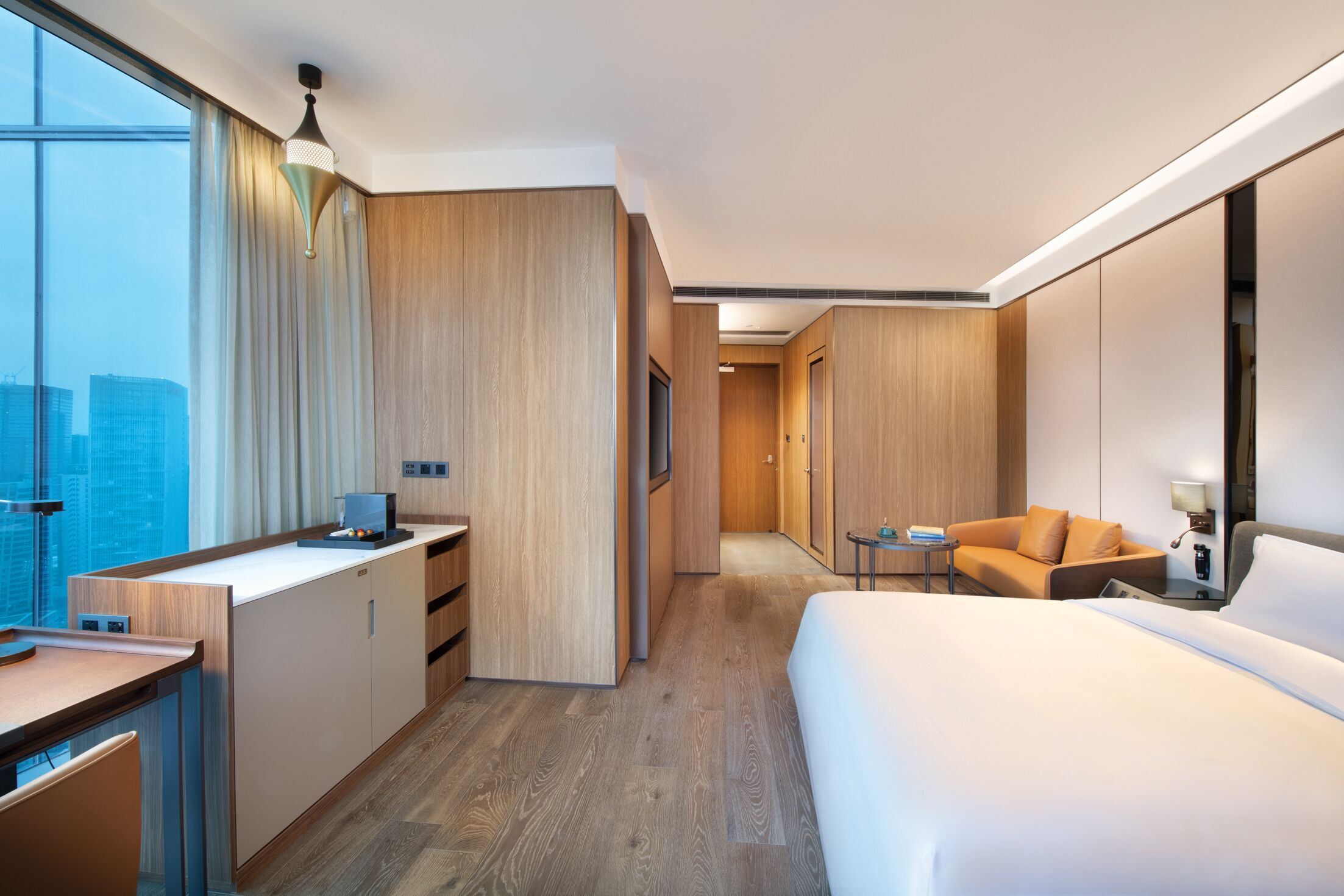 A modern hotel room with a large bed, a desk, a sofa, and a view of the city.