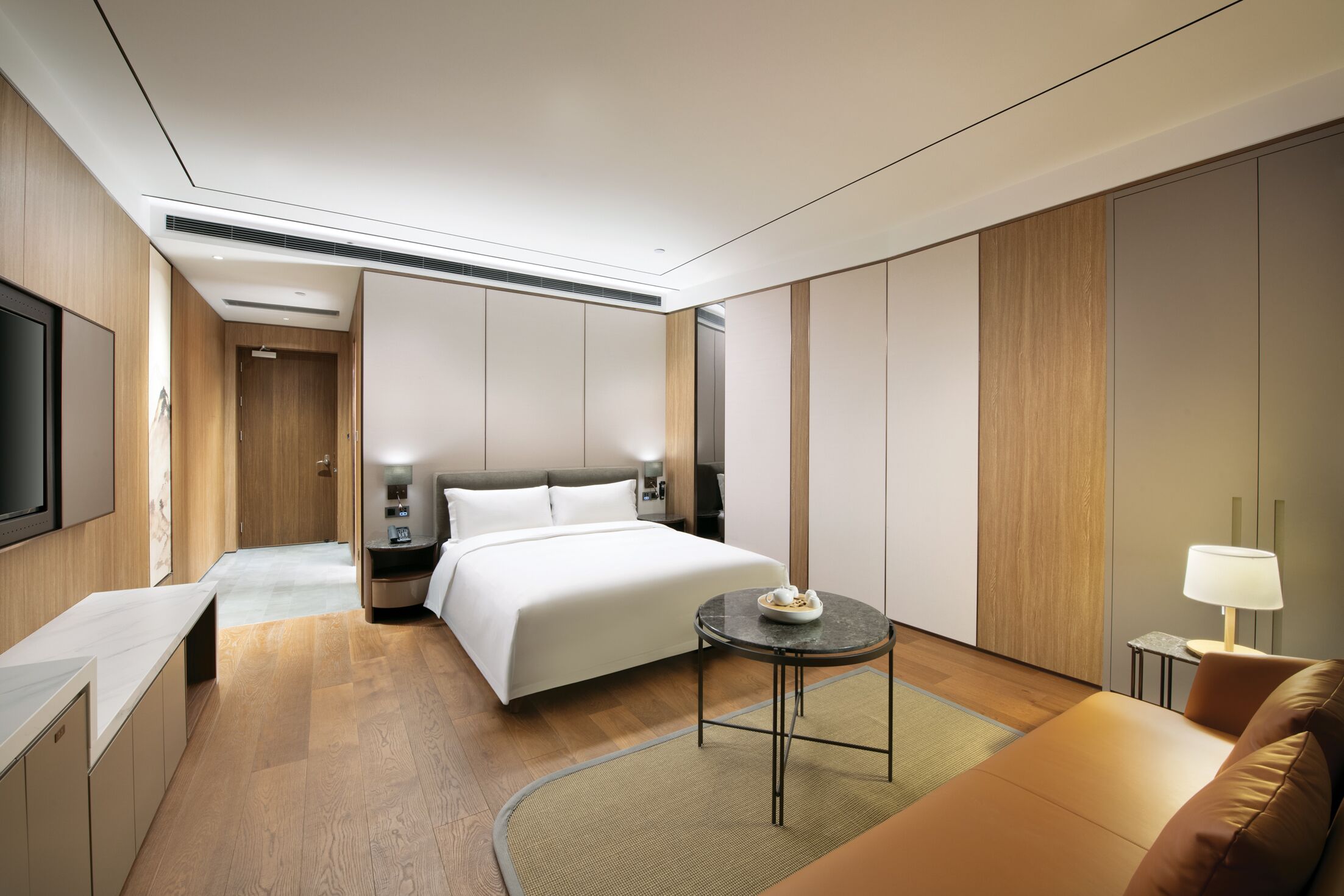 A modern hotel room with a bed, a small table, a sofa, a TV, and a lamp.