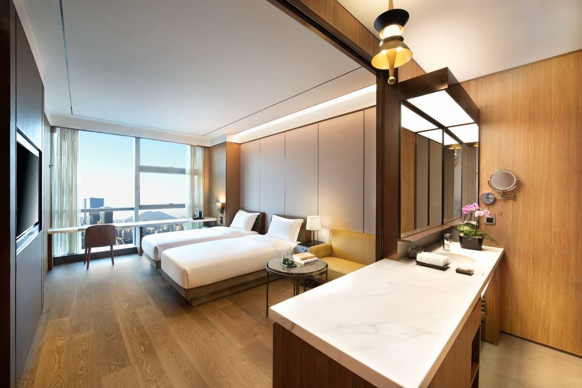 Habitación de hotel con dos camas, zona de estar, baño con espejo y lavamanos, y vista a la ciudad.