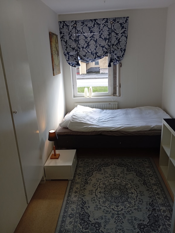 Ein kleines Schlafzimmer mit einem Einzelbett, einem Fenster mit gemusterten Vorhängen, einem Heizkörper, einem Nachttisch mit Lampe und einem Teppich mit Muster.