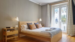 A modern bedroom with a wooden bed, bedside tables, a nightstand, and a view of the outside. - Fontana | The Rentals Collection (San Sebastián)